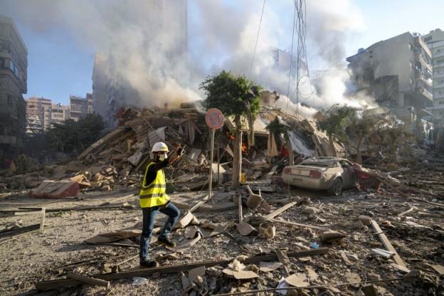 A man in a helmet, covering over his mouth, and reflective vest, gestures while standing on a road with much concrete debris, and a damaged vehicle.